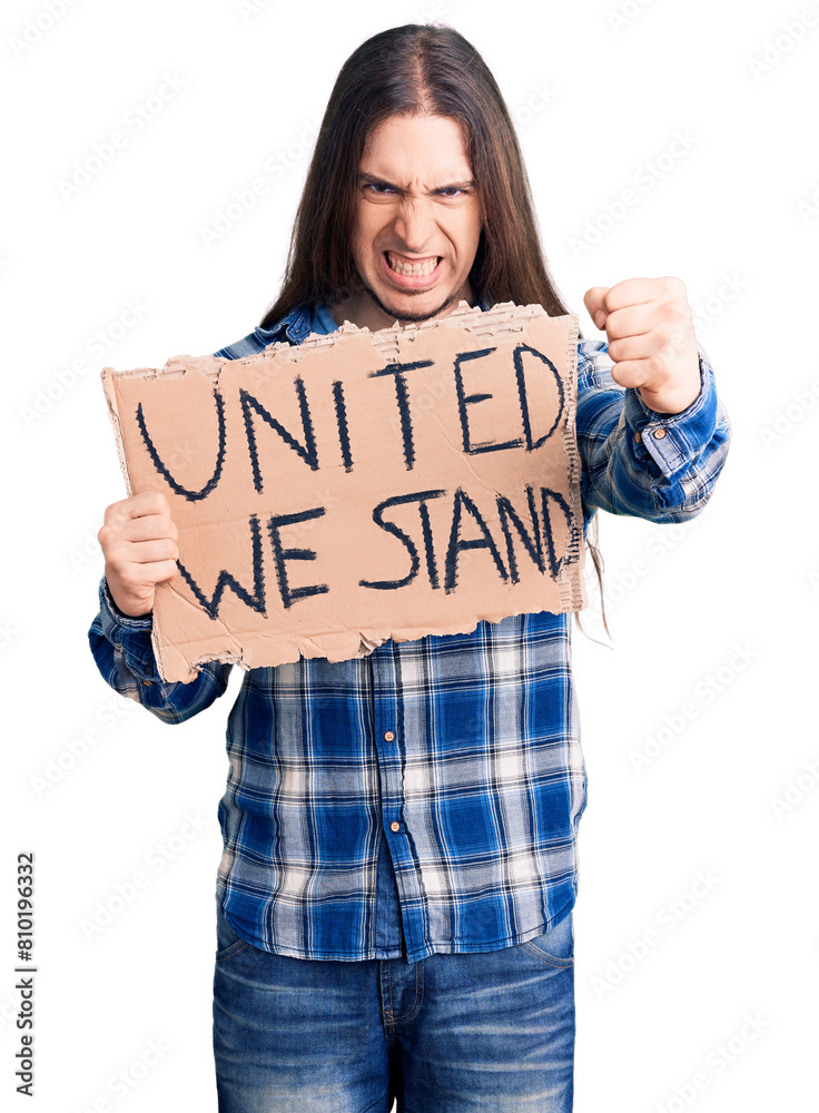 Young adult man with long hair holding united we stand banner annoyed and frustrated shouting with anger, yelling crazy with anger and hand raised Young adult man with long hair holding united we stand banner annoyed and frustrated shouting with anger, yelling crazy with anger and hand raised