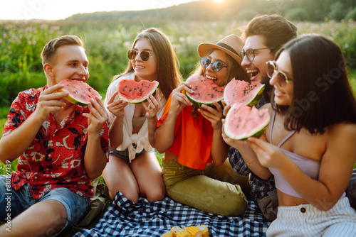 Cheerful friends eating fruits while sitting on the park. Young people having fun in summer vacation. Foods  travel  nature and picnic concept