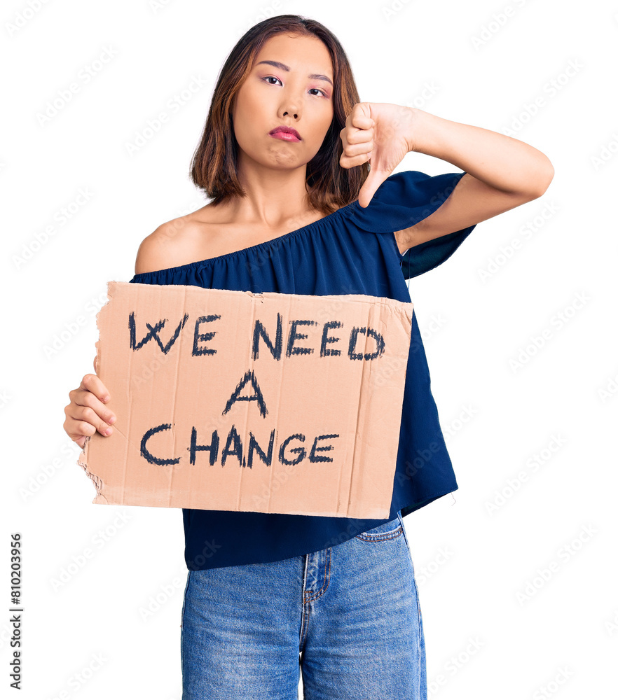 Young beautiful chinese girl holding we need a change banner with angry face, negative sign showing dislike with thumbs down, rejection concept Young beautiful chinese girl holding we need a change banner with angry face, negative sign showing dislike with thumbs down, rejection concept