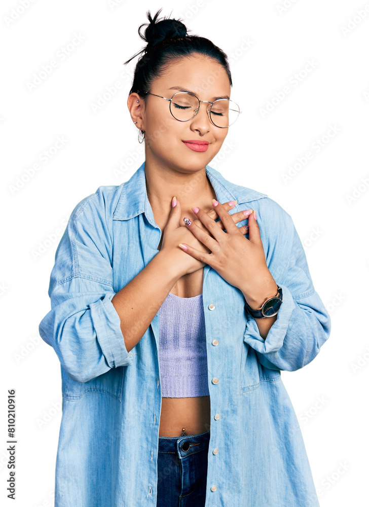 Beautiful hispanic woman wearing casual denim jacket and glasses smiling with hands on chest with closed eyes and grateful gesture on face. health concept.