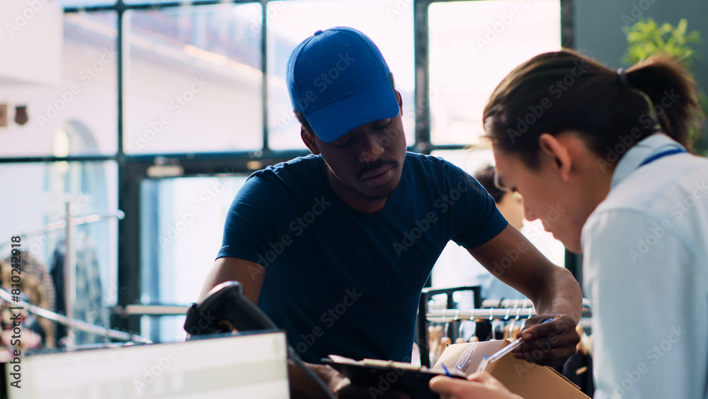 Asian manager signing delivery report, discussing shipping details with african american courier in clothing store. Employee preparing online orders for delivery in modern boutique Asian manager signing delivery report, discussing shipping details with african american courier in clothing store. Employee preparing online orders for delivery in modern boutique