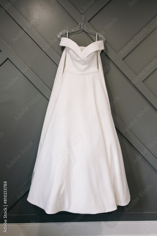 A white wedding dress hangs on a hanger on a gray wall in the bride's room. Beautiful dress of the bride on the wedding day. The bride's dress without decorations in a vertical photo A white wedding dress hangs on a hanger on a gray wall in the bride's room. Beautiful dress of the bride on the wedding day. The bride's dress without decorations in a vertical photo