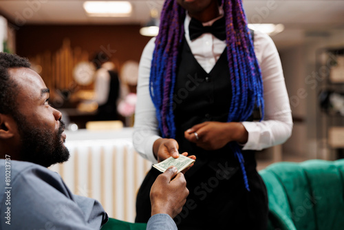 Bilde på lerret Young African American man hotel guest giving cash to waitress while relaxing in lobby, ordering food and drinks, tipping hospitality staff while resting at resort