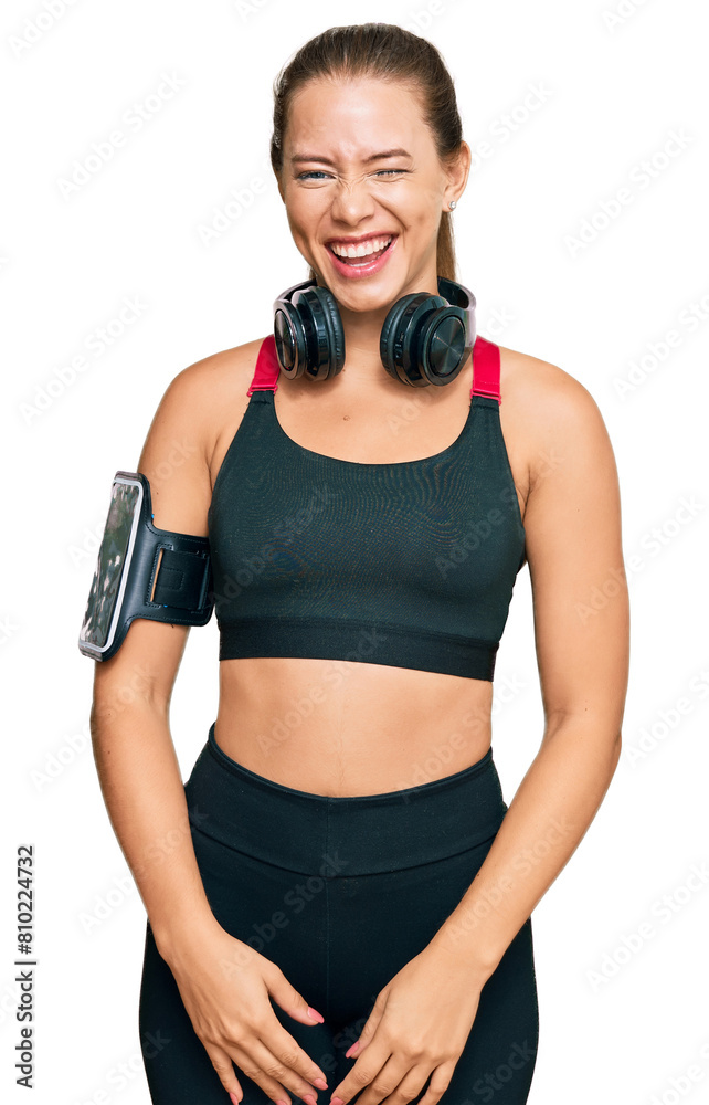 Beautiful blonde woman wearing gym clothes and using headphones winking looking at the camera with sexy expression, cheerful and happy face.