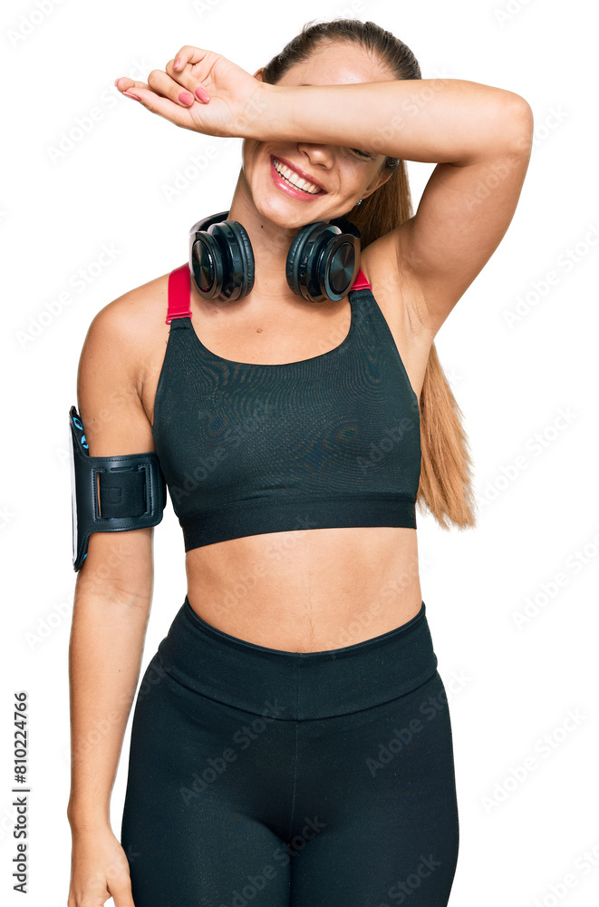 Beautiful blonde woman wearing gym clothes and using headphones covering eyes with arm smiling cheerful and funny. blind concept. Beautiful blonde woman wearing gym clothes and using headphones covering eyes with arm smiling cheerful and funny. blind concept.