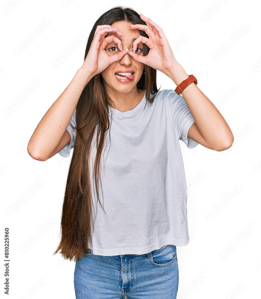 Young hispanic girl wearing casual white t shirt doing ok gesture like binoculars sticking tongue out, eyes looking through fingers. crazy expression. Young hispanic girl wearing casual white t shirt doing ok gesture like binoculars sticking tongue out, eyes looking through fingers. crazy expression.