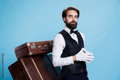 Professional hotel porter wears gloves posing over blue background, preparing to carry trolley bags and help guests. Young employee working as bellhop, tourism industry concept. photo