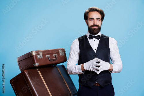 Young man hotel concierge with gloves helping guests to carry luggage and trolley bags, smiling with confidence in studio. Luxury doorman bellhop working in hospitality and tourism industry. photo