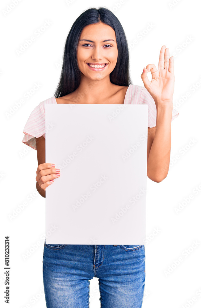 Young beautiful latin girl holding blank empty banner doing ok sign with fingers, smiling friendly gesturing excellent symbol Young beautiful latin girl holding blank empty banner doing ok sign with fingers, smiling friendly gesturing excellent symbol