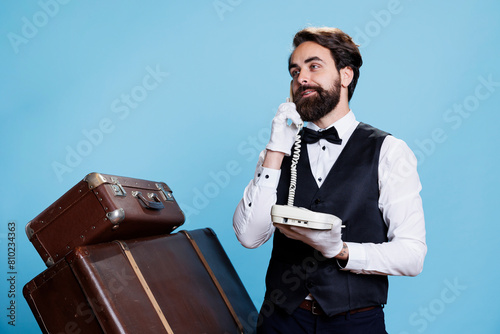 Stylish bellboy answers landline phone in studio, talking to hotel guests about accommodation. Skilled confident doorman using telephone with cord to take calls, vintage luxurious job. photo