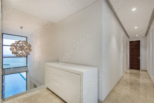 The hallway in the villa has white walls  beige floor tiles and large windows..