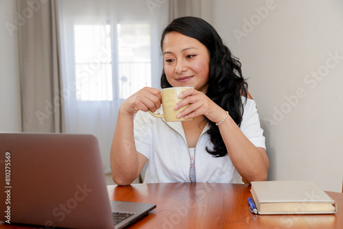 Hispanic adult woman drinking coffee while working on her computer - Happy woman at home watching her computer screen - Remote work - Work video call - Woman studying at home
