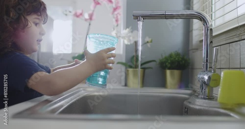 A boy carefully fills a clear glass from a stainless steel sink tap, natural daylight illuminating the cozy kitchen scene.