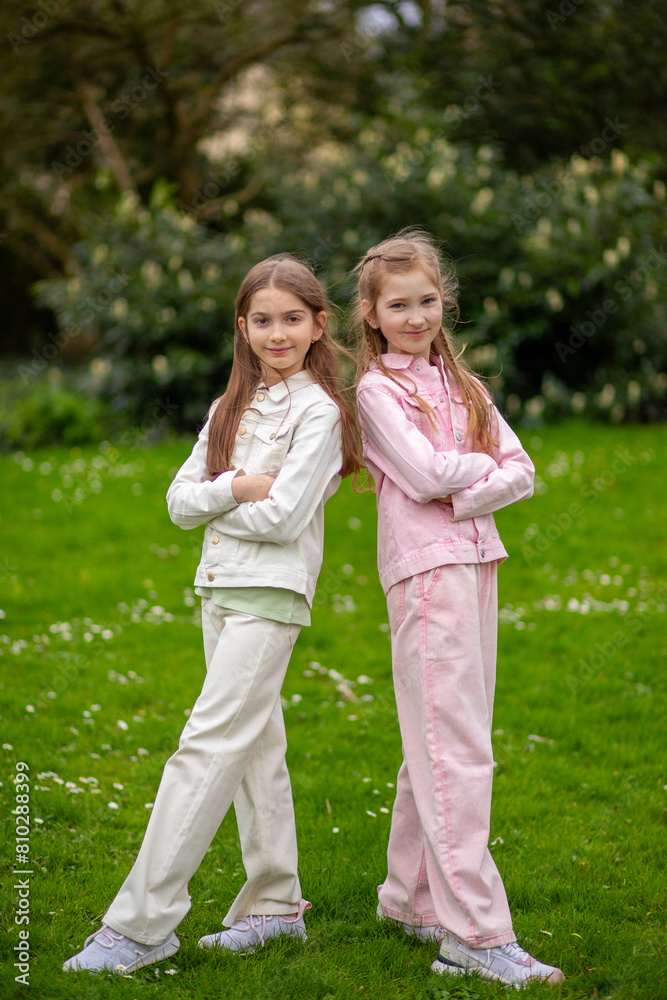 Two friendly girls in light-toned clothing with smiles enjoying a spring day in the park, standing on green grass. Two friendly girls in light-toned clothing with smiles enjoying a spring day in the park, standing on green grass.