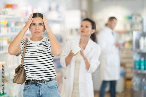 Young girl feeling pain standing with her back to female pharmacist offering remedy to her in drugstore