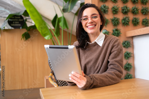 Woman holding tablet computer sitting at her desk smile and looking camera. Female freelancer person studying or working online on touch pad. Modern technologies and communication.