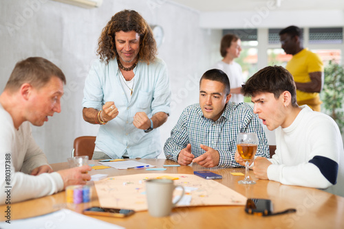 Expressive cheerful man with long wavy hair enthusiastically playing board game with male friends of different nationalities during bachelor party