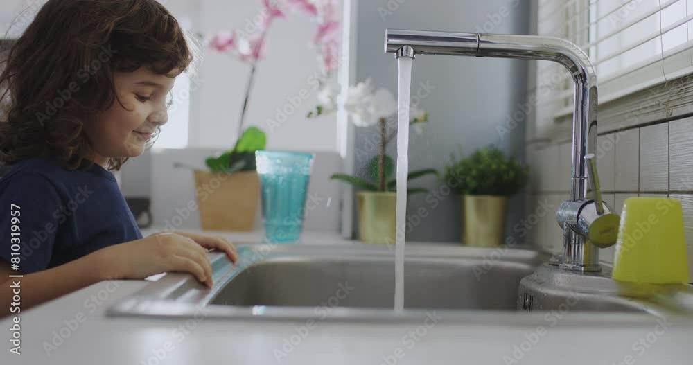 A curious young boy is closely observing the stream of water flowing from a modern kitchen faucet, with a focus on the clear, running water. A curious young boy is closely observing the stream of water flowing from a modern kitchen faucet, with a focus on the clear, running water.