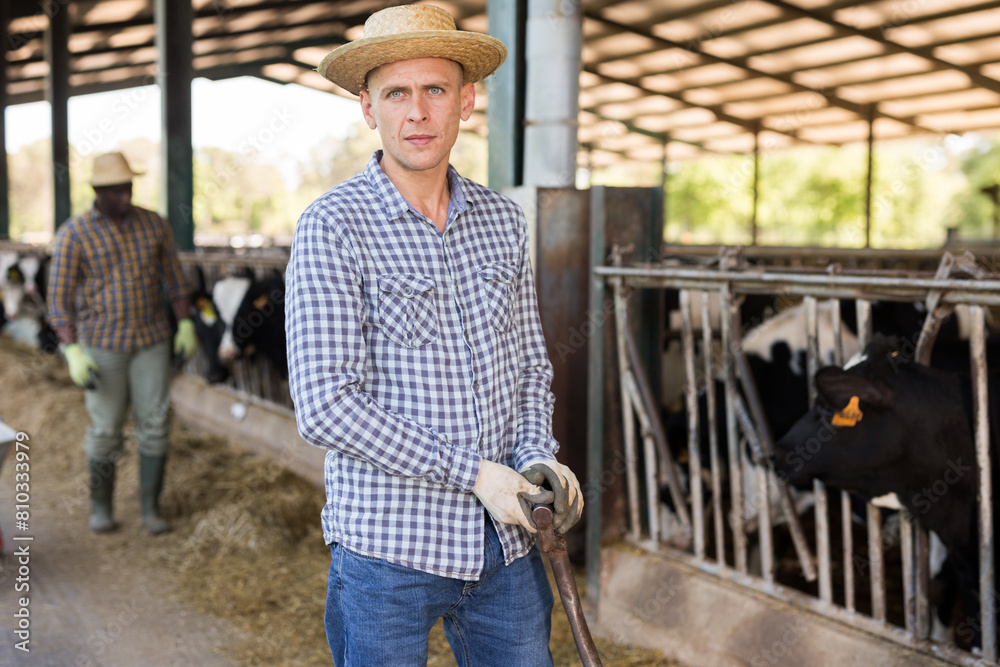 Portrait of a confident young male farmer standing in a cowshed with a shovel in his hand. Close-up portrait