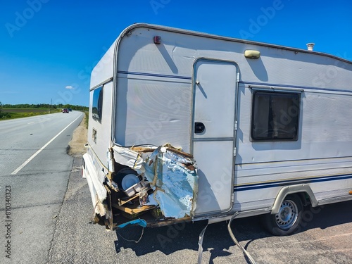 Vintage white travel trailer with retro design and visible damage to the back left corner resulting from a recent accident on the side of the road under clear blue sky on a bright sunny day