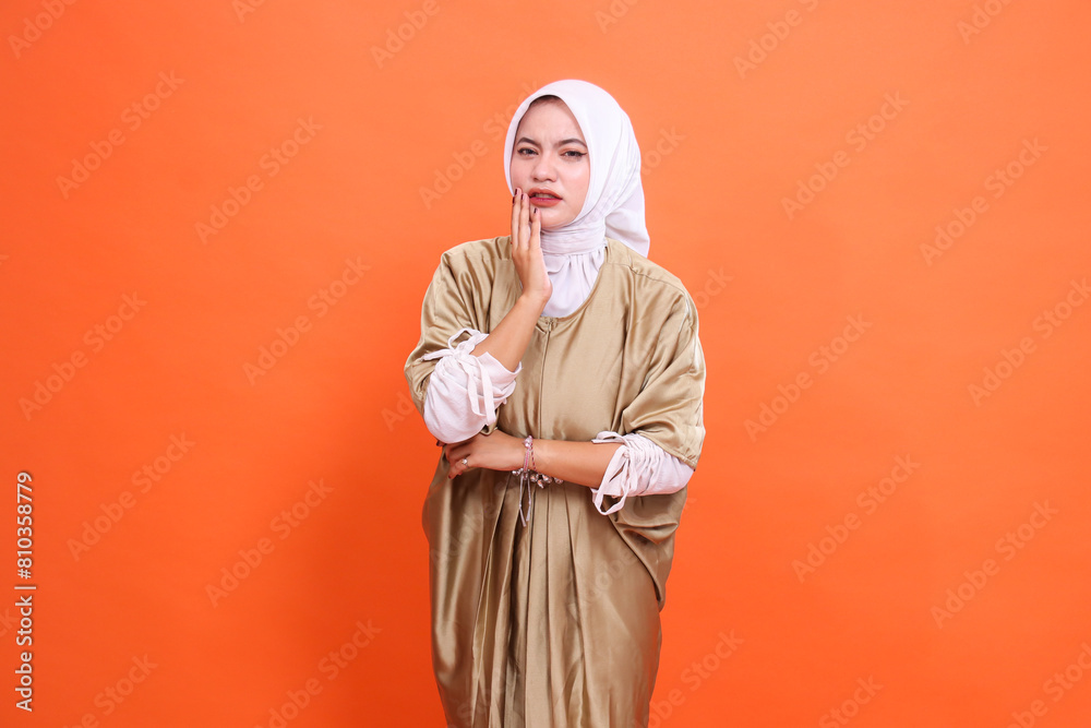 The expression of a beautiful adult Asian woman is sad, her teeth ache, holding her cheek, wearing a Muslim dress, kaftan, hijab, isolated on an orange background. Concept of health care, education The expression of a beautiful adult Asian woman is sad, her teeth ache, holding her cheek, wearing a Muslim dress, kaftan, hijab, isolated on an orange background. Concept of health care, education