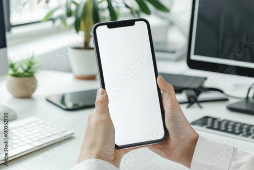 woman holding phone mockup with two hands, against the background of an office table, keyboard, computer and houseplants