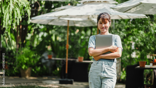 Female freelancer stands with her laptop in a lush garden, representing a serene remote work setting.