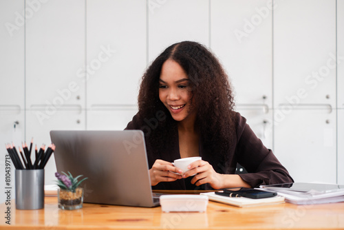 Smiling businesswoman enjoys a cup of coffee while working on her laptop, exuding a relaxed yet productive office atmosphere. © Mongta Studio