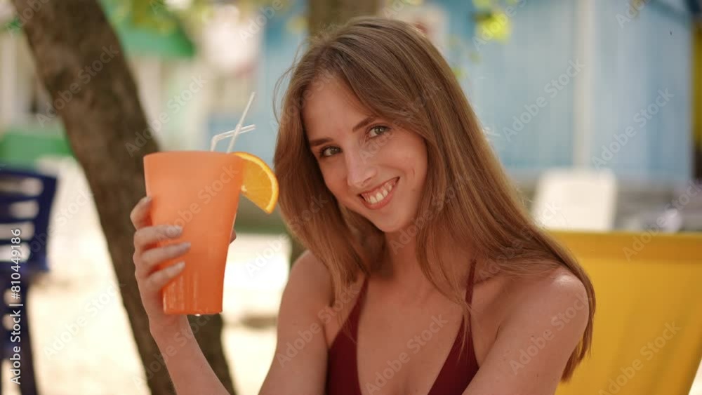 Slow motion. Close-up. Cute girl smiling looking at the camera and holding a cocktail with a cocktail straw in her hand while sitting on a beach chair on a sunny summer day outdoors Slow motion. Close-up. Cute girl smiling looking at the camera and holding a cocktail with a cocktail straw in her hand while sitting on a beach chair on a sunny summer day outdoors