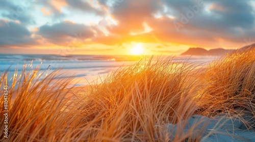 Golden sunrise illuminates beachgrass on sandy shore with distant mountains and vibrant sky