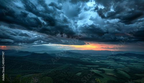 A panoramic view of the sky at sunset, with dark storm clouds rolling in over distant hills. The sun's rays pierce through these thick grey and blue hues, casting long shadows on the landscape below. 