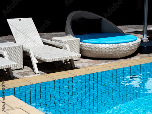 Empty white modern rattan sunbeds chair, and waterproof round lie bed with blue seat cushion for guests at the poolside in hotel resort with nobody. Summer swimming pool relaxation background concept.