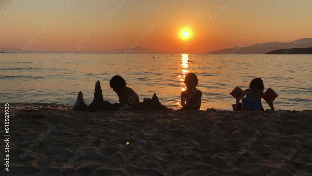 A group of people, including kids, sitting on the sandy beach. The sun is setting in the background, creating a warm glow over the scene. A group of people, including kids, sitting on the sandy beach. The sun is setting in the background, creating a warm glow over the scene.