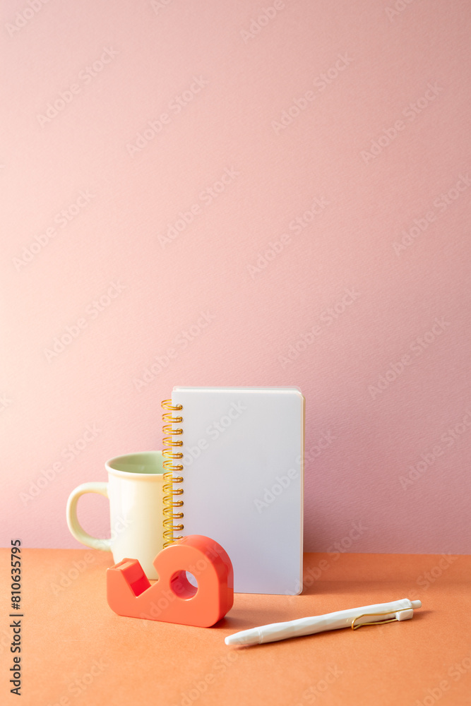 Workspace office desk with notebook, mug cup, pen, tape dispenser. pink orange background