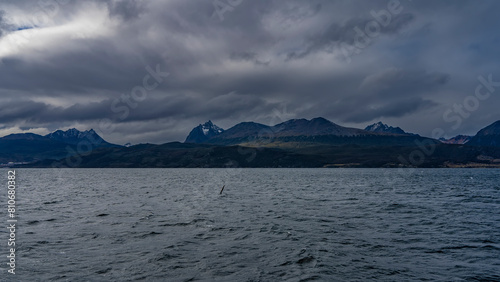 The picturesque snow-capped mountain range is visible from the ocean. Dark clouds in the sky. Seabirds fly over the water. Tierra del Fuego Archipelago. The Beagle Channel. The Andes.