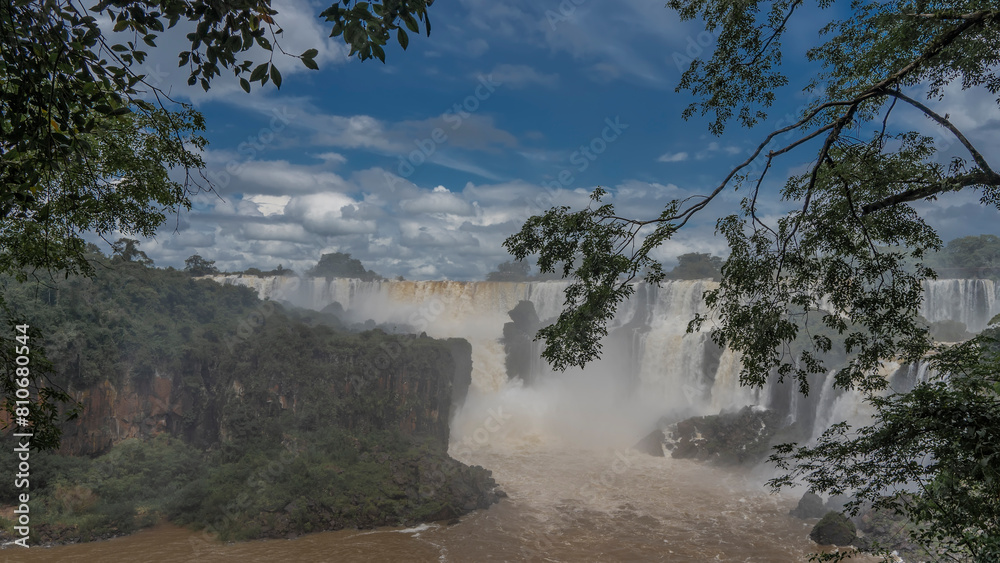 Beautiful tropical waterfall landscape. Foaming streams fall from a ledge in the riverbed. Splashes. Green vegetation on the rocks. Tree branches against a blue sky and clouds. Iguazu Falls. Argentina