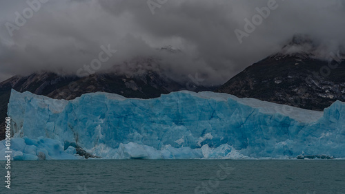A block of blue ice rises like a wall above a glacial lake. Coastal mountains in clouds and fog. Icebergs float in glacial water. Perito moreno glacier. El Calafate. Argentina. Lago Argentino. 
