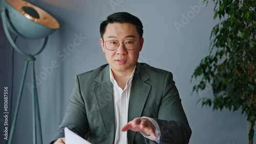 View from modern laptop camera of talkative japanese male gesturing while holding paper documents on background of designer cabinet interior. Handsome worker taking part in online conversation. photo
