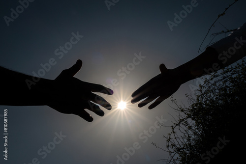 Silhueta de duas mão em que a mão feminina em meio a silvas e às adversidades da vida pede ajuda à mão masculina com o sol como fundo significando a esperança photo