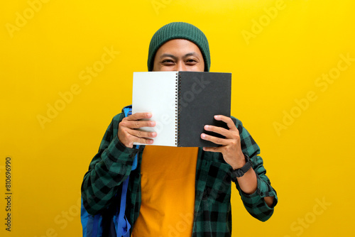 A happy and confident young Asian male student, dressed in a beanie hat and casual clothes, smiles at the camera while partially covering half of his face with a notebook isolated on yellow background