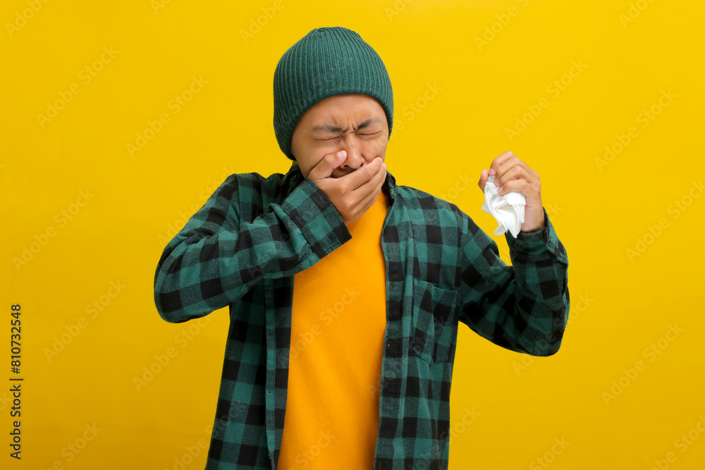 Young Asian man, wearing a beanie hat and casual shirt, is holding a napkin tissue while experiencing throat pain and dry coughing, suggesting discomfort, isolated on a yellow background. Young Asian man, wearing a beanie hat and casual shirt, is holding a napkin tissue while experiencing throat pain and dry coughing, suggesting discomfort, isolated on a yellow background.