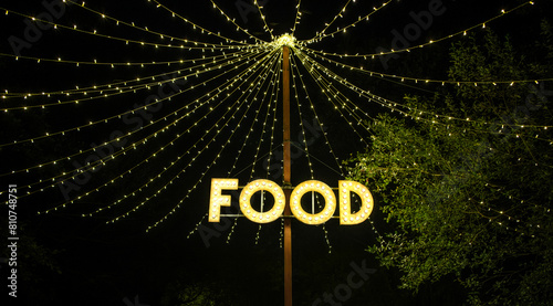 Signboard with word food made with light bulbs hung on a wooden post under light garlands located in the middle of treetops at night. Concept of food and drink zone on outdoors summer festival.