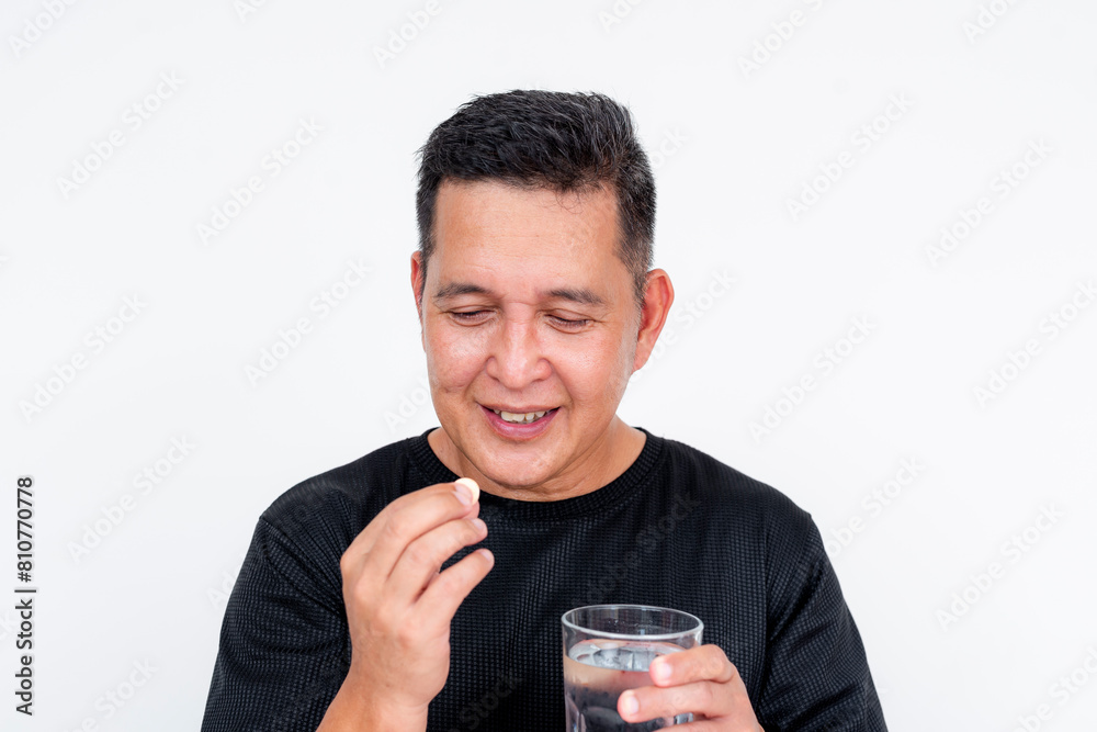 A middle-aged Asian man smiles as he takes a sodium bicarbonate antacid tablet, holding a glass of water, isolated on a white background. A middle-aged Asian man smiles as he takes a sodium bicarbonate antacid tablet, holding a glass of water, isolated on a white background.