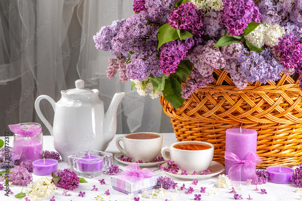 A basket of purple flowers sits on a table with a white tea set and candles A basket of purple flowers sits on a table with a white tea set and candles