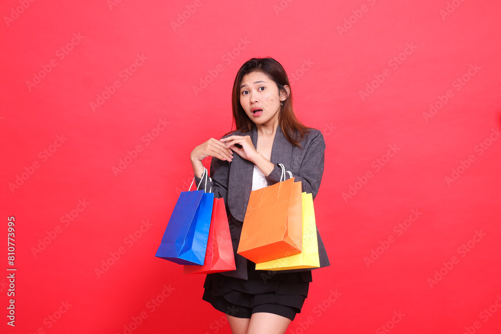 Expression of shocked indonesia office woman carrying shopping paper bags with her hands in front of her shoulders wearing a formal jacket on a red background. for commercial, lifestyle and sales Expression of shocked indonesia office woman carrying shopping paper bags with her hands in front of her shoulders wearing a formal jacket on a red background. for commercial, lifestyle and sales
