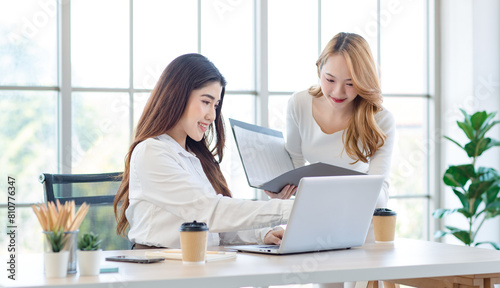 Asian young pretty female businesswoman team smiling discussing project on laptop standing and sitting at table in office. Two entrepreneurs colleagues of professional business people working together