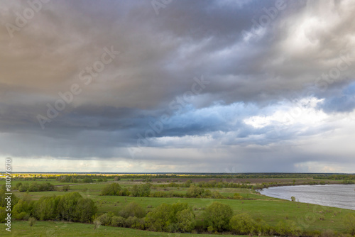 A tranquil rural landscape under a dramatic sky  where storm clouds gather above lush green fields and a distant river  casting a play of light and shadow across the scene