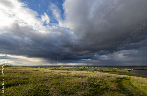 A tranquil rural landscape under a dramatic sky, where rolling hills of green grass meet a distant horizon under a mix of dark storm clouds and sunlight.