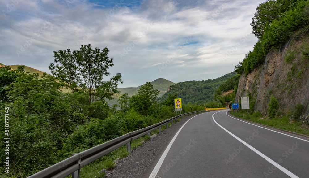 A winding mountain road with a guardrail on the left, bordered by lush green trees and vegetation. Yellow caution signs and white road markings indicate a driving perspective on an overcast day A winding mountain road with a guardrail on the left, bordered by lush green trees and vegetation. Yellow caution signs and white road markings indicate a driving perspective on an overcast day
