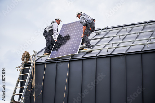 Electricians installing solar panel system on roof of house. Men workers in helmets carrying photovoltaic solar module outdoors. Concept of alternative and renewable energy.